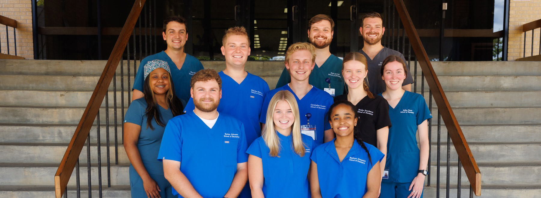 Group of healthcare trainees wearing blue and black scrubs standing together on indoor stairs in a campus building.